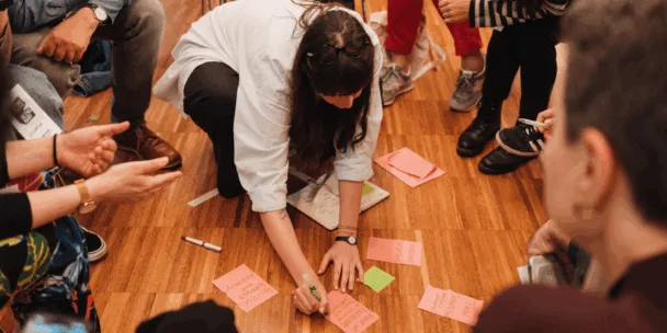 Un grupo de personas se sienta en círculo sobre un suelo de madera, participando en un taller. En el centro, una mujer de pelo largo y oscuro, con gafas, se arrodilla y escribe en tarjetas de papel de color naranja y rosa brillante. Otros participantes sostienen bolígrafos y gesticulan mientras conversan. Algunas tarjetas en el suelo están etiquetadas con palabras escritas a mano.