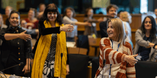 A group of people standing in a European Parliament conference room engage in a group activity, smiling and extending one arm forward while placing the other arm across their chest. The mood is warm and lighthearted. Participants of various ages and backgrounds appear to be following movement and dancing instructions.
