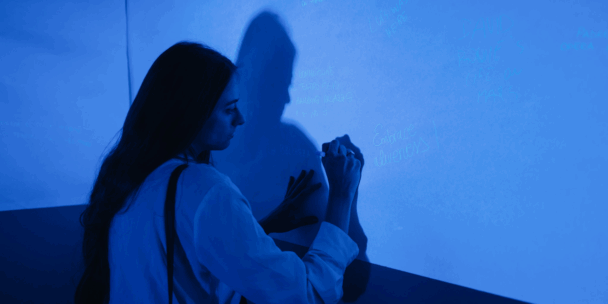 A woman with long hair, wearing a light-colored shirt, writes on a large blue wall with a pen under blue lighting. Her shadow is cast clearly on the wall. Faint handwritten messages and names are visible across the surface.