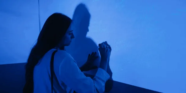 Una mujer de cabello largo, vestida con una camisa clara, escribe con un bolígrafo en una gran pared azul bajo una luz azul. Su sombra se proyecta nítidamente sobre la pared. Se ven tenues mensajes y nombres manuscritos sobre la superficie.