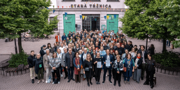 A large group of over 100 people pose for a photo outside the historic Stará Tržnica building in Bratislava, Slovakia. The participants stand under colorful bunting and between two leafy trees, smiling toward the camera. Behind them, two large green banners announce the cultural conference programme on 29–30 May 2025. At the front, several people hold printed documents.