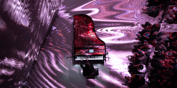 An overhead view of a pianist performing on a grand piano with its lid open, revealing red-toned inner mechanics. The floor and surrounding walls are covered in swirling, psychedelic purple and white light projections. A large audience sits to the right, attentively watching the performance, immersed in the vivid atmosphere of the space. The setting evokes a futuristic, multisensory arts experience.