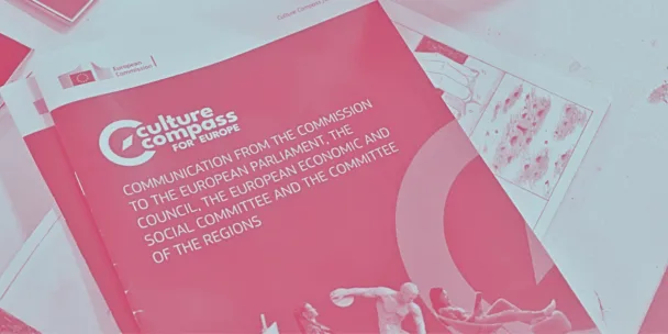 Una fotografía en tonos rosados ​​de una mesa con un folleto titulado Brújula Cultural para Europa —una publicación de la UE sobre política cultural— junto a notas manuscritas, páginas ilustradas y una caja que dice “ACTÚA” con un código QR.