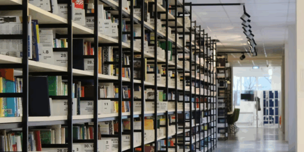 Bookshelves take up two thirds of the photo, as part of a public library with many different types of books and a long, well-lit corridor.