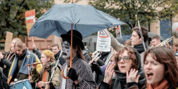 Fotografia pouličného protestu alebo verejnej demonštrácie. V strede je osoba v bielo-čiernom pantomímickom farbe na tvár, s cylindrom a kockovanou košeľou, ktorá drží čierny dáždnik. Okolo nich je aktívnych niekoľko ďalších ľudí: jeden hrá na modrý bubon, druhý na flautu a niekoľko ďalších zrejme kričí alebo skanduje. Pozadie je mierne rozmazané stromami a mestskými budovami.