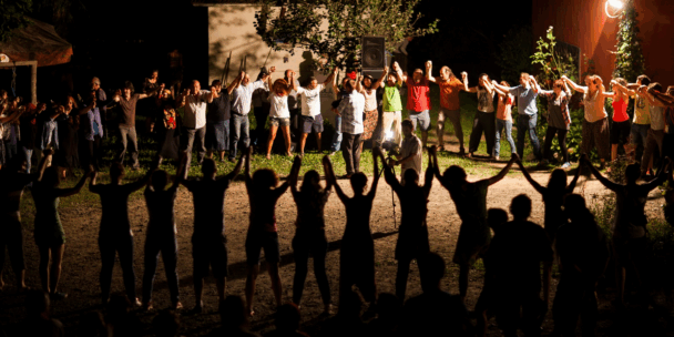 A large group of people form a wide circle, holding hands in solidarity under warm lighting in an outdoor nighttime setting. Some stand in shadow while others are brightly illuminated, creating a powerful silhouette effect. Trees, a red building, and a speaker are visible in the background, adding to the sense of a communal, cultural gathering.