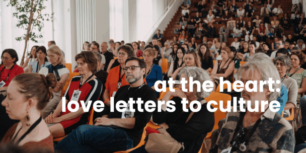 A large audience sits closely together in a bright auditorium with wooden tiered seating. Many people wear conference badges on lanyards and appear reflective, some with eyes closed as if listening intently or participating in a quiet moment. The crowd is diverse in age and gender. White overlaid text across the image reads: “at the heart: love letters to culture.”