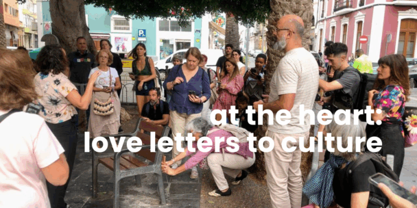A group of around fifteen people gather outdoors under leafy trees on a city street. A woman gestures while speaking to the group, who listen attentively; several hold smartphones. One older woman crouches near a wooden bench, appearing to examine or adjust something at its base. The setting includes palm trees, parked cars, pastel-coloured buildings, and street signs in the background. White overlaid text across the image reads: “at the heart: love letters to culture.”