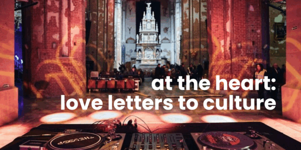 Interior of a large historic church or cathedral repurposed for a cultural event. Brick columns and high vaulted walls are illuminated with warm orange and yellow light projections. At the far end, an ornate white altar stands beneath tall windows. In the foreground, DJ equipment and turntables sit on a table facing the open space. Small groups of people gather in the middle distance. Overlaid in large white text: “at the heart: love letters to culture.”