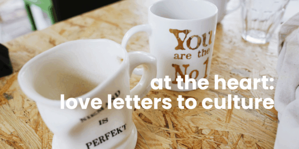 A close-up of two ceramic coffee mugs on a light wooden table with a natural chipboard texture. One mug in the foreground is slightly stained inside; the other, behind it, has gold lettering partially visible. A glass of water and a folded white cloth sit to the right. Overlaid in large white text across the image: “at the heart: love letters to culture.”