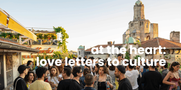 A large group of people gather on a rooftop terrace at sunset, socialising and talking in small clusters. The setting features greenery, café awnings, and historic buildings with domes and a tall tower in the background. The light is warm and golden. White overlaid text across the image reads: “at the heart: love letters to culture.”