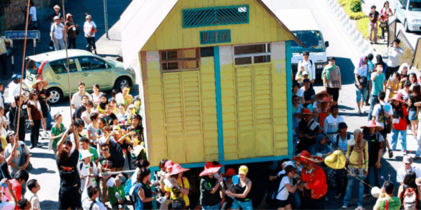 A large, yellow and green wooden house is being carried through a crowded street by a group of people, some of whom are wearing colorful hats and uniforms. The street is filled with many onlookers, some taking photos and others watching the unusual procession near parked cars and a white van. The bright, high-angle shot captures a lively, communal event taking place outdoors in a sunny environment.