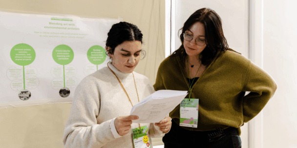 Two women wearing event lanyards stand together in an indoor setting, focused on reviewing a printed document held by the woman on the left. In the background, a poster displays green circular diagrams and text about climate change initiatives. Both women appear engaged in a professional discussion or collaboration during a workshop or conference.