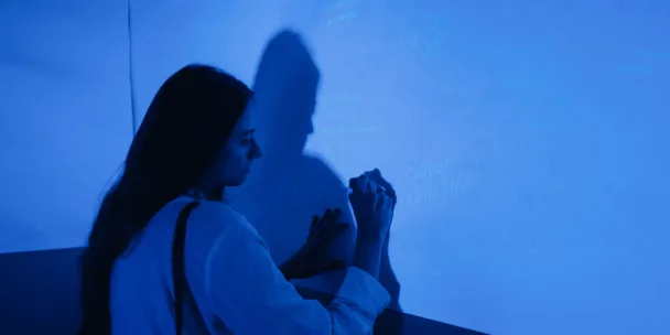 A woman with long dark hair is seen in profile as she writes on a wall illuminated by a vibrant blue light. Her silhouette is cast as a large shadow against the wall, which is covered in various handwritten words and phrases like "Embrace aliveness." The scene has a moody, artistic atmosphere with high contrast between the dark foreground and the glowing blue surface.