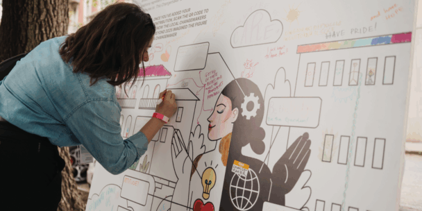 A person in a blue denim shirt is seen from the side, drawing with a marker on a large white board outdoors next to a tree. The board features a collaborative mural with illustrations of buildings, a central figure with a gear in their hair, and various handwritten messages. The artwork includes colorful accents in pink, yellow, and red, creating a vibrant scene of community engagement and creativity.