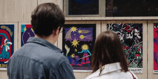 Two people are seen from behind, looking at a series of colorful, stylized posters displayed on a light-colored exterior wall. The central poster features a dark, celestial theme with yellow star-like characters, a sword-wielding figure, and a wolf. To the right, another vibrant poster depicts a dense, psychedelic forest scene filled with intricate patterns and small animal-like figures.