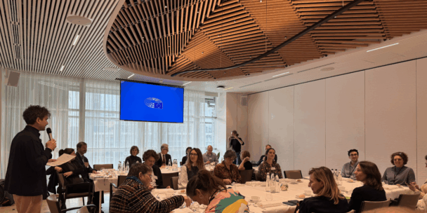A meeting room in the European Parliament set up for a policy lunch, with participants seated around round tables while a speaker stands at the left holding a microphone and notes. A large blue screen with the European Parliament logo hangs in front of tall windows, and the room features a sculptural wooden ceiling. Attendees listen, take notes, and talk quietly around tables set with water bottles, glasses, and coffee cups.