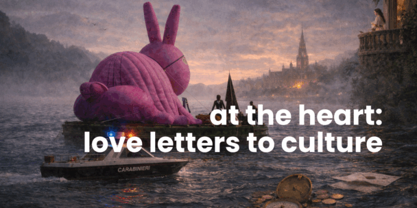 A surreal, cinematic scene at dusk shows a large pink rabbit sculpture floating on a raft in a misty river. Two figures stand on the raft beside it. In the foreground, a small patrol boat labeled “Carabinieri” moves across the water with blue and red lights flashing. On the right, a woman stands on a stone balcony overlooking the scene. In the distance, a historic city skyline with a tall cathedral spire glows warmly under a cloudy evening sky. Overlaid in large white text: “at the heart: love letters to culture.”