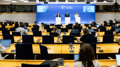 A wide shot from the back of a large press room shows three people standing at white podiums in front of a blue backdrop labeled "European Parliament." Numerous journalists are seated at long yellow desks in the foreground, facing the speakers with laptops open. The room is modern and brightly lit by rows of long, parallel ceiling lights.