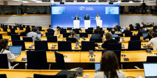 A wide shot from the back of a large press room shows three people standing at white podiums in front of a blue backdrop labeled "European Parliament." Numerous journalists are seated at long yellow desks in the foreground, facing the speakers with laptops open. The room is modern and brightly lit by rows of long, parallel ceiling lights.