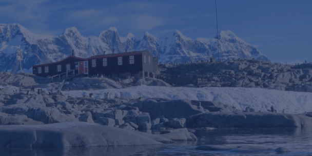 A remote research station with a long, dark building and a red door sits on a rocky, snow-dusted shoreline. In the background, sharp, majestic snow-capped mountain peaks rise under a soft sky. Numerous penguins are scattered across the rocky foreground near the water's edge, all viewed through a cool, blue-tinted overlay.