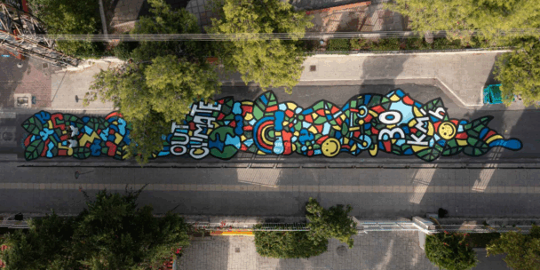 An aerial, top-down view shows a long, vibrant street mural painted directly onto the asphalt between two sidewalks. The colorful, mosaic-style artwork features bold black outlines and includes the phrases "OUR CLIMATE" and "30 KM/h" alongside symbols like a globe, a rainbow, and smiley faces. Lush green trees and urban architecture border the street, framing the artistic intervention within the city landscape.