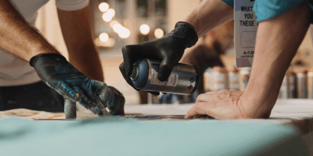A close-up shot focuses on the hands of two people working over a table, with one person wearing a black gloved hand holding a silver and blue spray paint can aimed at a surface. The other person's hand, also in a black glove, is covered in blue and green paint residue as they guide or hold the artwork in place. In the blurred background, other spray cans and a sign are visible under warm, out-of-focus lighting.