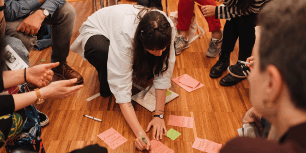 A close-up, high-angle view captures a woman crouching on a wooden floor, writing on pink sticky notes with a green marker. She is surrounded by a group of people sitting or kneeling in a circle, some of whom are holding pens and notebooks. Scattered around her on the floor are an open notebook, a white pen, and multiple pink and green sticky notes containing handwritten text. The group is clearly collaborating and working on something together.
