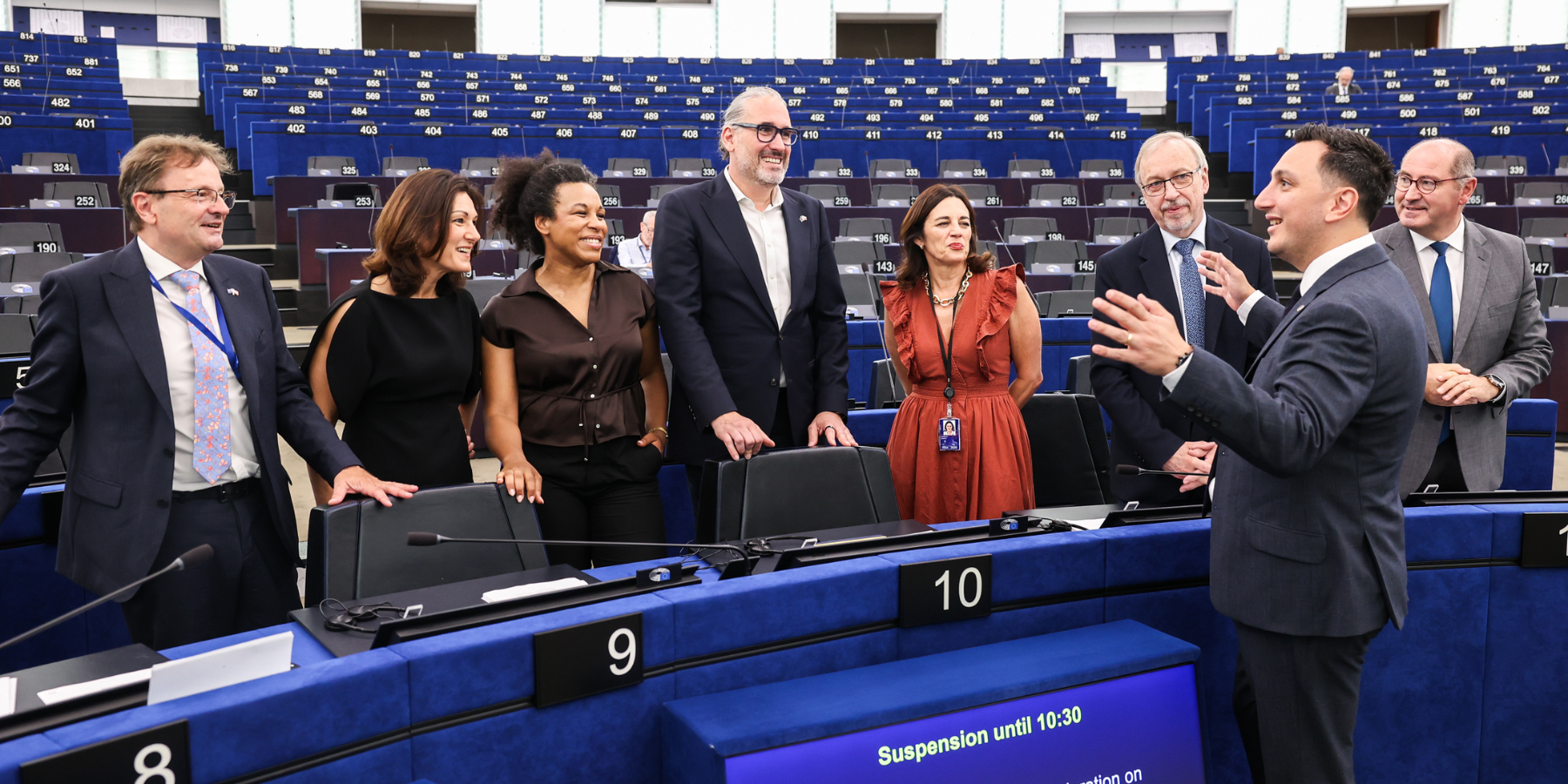 A group of European Union politicians and officials are standing together inside the European Parliament hemicycle, with rows of empty blue parliamentary seating rising behind them. One politician on the right is actively gesturing and speaking to the group, who are listening and smiling. In the foreground, digital screens built into the desks display legislative scheduling info,