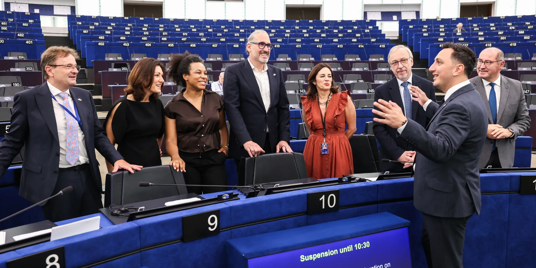 Eine Gruppe von EU-Politikern und Beamten steht im Halbkreis des Europäischen Parlaments, hinter ihnen erheben sich Reihen leerer blauer Parlamentssitze. Ein Politiker rechts gestikuliert lebhaft und spricht zu der Gruppe, die ihm zuhört und lächelt. Im Vordergrund zeigen in die Tische integrierte digitale Bildschirme Informationen zum Sitzungsplan an.