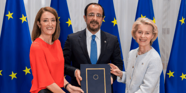 European leaders Roberta Metsola, Nikos Christodoulides, and Ursula von der Leyen stand side-by-side smiling while jointly holding a dark blue document folder embossed with the European Union stars. They are formally dressed and positioned in front of a backdrop featuring several large European Union flags.