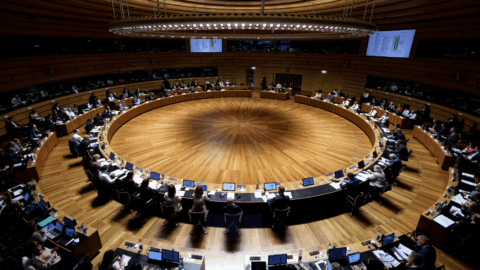 An elevated, high-angle view of the image shows a large, circular conference hall with wooden flooring and multiple tiers of seating where numerous people are seated at desks equipped with monitors. A giant, ring-shaped light fixture hangs from the concentric wooden ceiling directly over the center of the room. Digital display screens are mounted on the upper wooden walls, overlooking the formal meeting of the European Council.