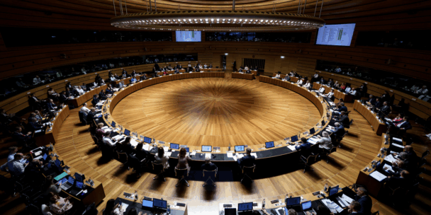 An elevated, high-angle view of the image shows a large, circular conference hall with wooden flooring and multiple tiers of seating where numerous people are seated at desks equipped with monitors. A giant, ring-shaped light fixture hangs from the concentric wooden ceiling directly over the center of the room. Digital display screens are mounted on the upper wooden walls, overlooking the formal meeting of the European Council.