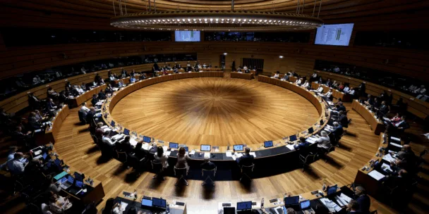 Une vue aérienne de l'image révèle une vaste salle de conférence circulaire au parquet en bois et aux multiples gradins où de nombreuses personnes sont assises à des bureaux équipés d'écrans. Un imposant luminaire annulaire est suspendu au plafond en bois concentrique, directement au-dessus du centre de la salle. Des écrans numériques sont fixés aux murs supérieurs en bois, donnant sur la réunion officielle du Conseil européen.
