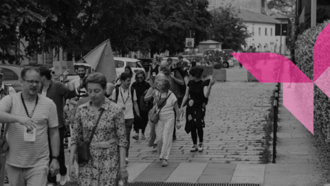 A black-and-white photograph shows a diverse group of people walking down a paved, cobblestone European street or pedestrian walkway lined with trees and buildings. Some individuals are talking and smiling, and a person in the background holds up a light-colored flag. A stylized, bright pink geometric butterfly graphic is overlaid on the right side of the image, contrasting with the monochrome scene.