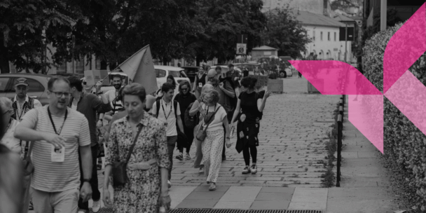 A black-and-white photograph shows a diverse group of people walking down a paved, cobblestone European street or pedestrian walkway lined with trees and buildings. Some individuals are talking and smiling, and a person in the background holds up a light-colored flag. A stylized, bright pink geometric butterfly graphic is overlaid on the right side of the image, contrasting with the monochrome scene.