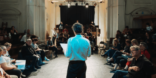 A man with curly dark hair, seen from behind and lit in blue-toned light, stands in the center of a large room reading from a piece of paper held in his hand. He faces a seated audience of conference attendees wearing name badges, arranged in a wide circle around him in what appears to be a converted theater or hall with ornate architectural columns and a stage with lighting rigs in the background. Papers, phones, and bags are scattered among the seated participants, who watch and listen attentively.