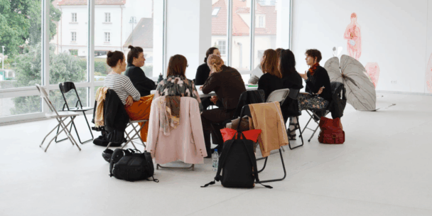 A group of about nine people sits in a circle of chairs having a discussion in a bright, white-walled gallery space with large windows overlooking historic rooftops and trees outside. Bags, backpacks, and jackets are draped over several of the empty chair backs and piled on the floor. In the background stands a large artistic sculpture/installation resembling anatomical figures and butterfly-like translucent wings mounted on the wall.