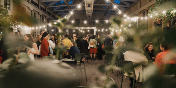 A group of people are in a bar that is well lit, standing speaking with one another across diverse tables.