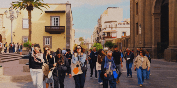 A group of people walking through a historic, stone-paved European plaza lined with warm-colored buildings, palm trees, and a large stone church on the right.