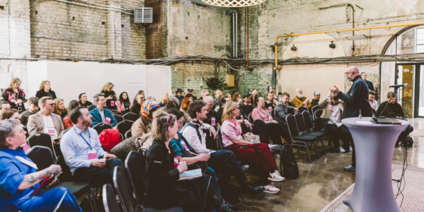 A large seated audience wearing pink conference lanyards listens to a man in a dark jacket speaking with animated hand gestures near a standing podium with a laptop and microphone. The event takes place in an industrial-style venue with exposed brick walls, visible pipes and cables, and a decorative lattice-shaped light fixture hanging from the ceiling. An open doorway on the right lets in natural light from outside.
