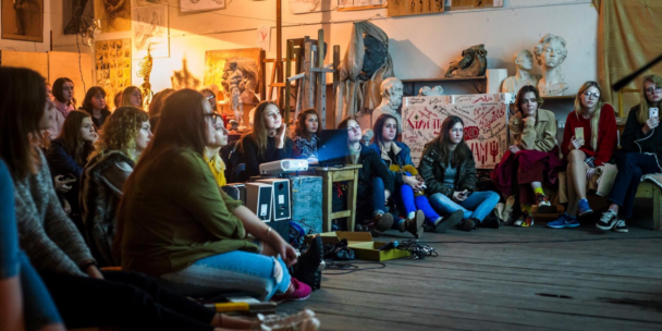 A group of students sits attentive on the floor and chairs inside a workshop or art studio space, looking toward a presentation off-camera. A digital projector sits on a small wooden table in the center, casting a light beam across the room alongside speakers placed in the foreground. Classical busts, statues, and a wall covered in graffiti and handwritten text fill the background behind the audience. The overall atmosphere feels warm, collaborative, and creative.