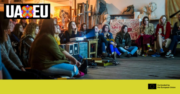 Young women sit close together on the floor of an art studio, watching a presentation. UA EU logo. Co-funded by the European Union.