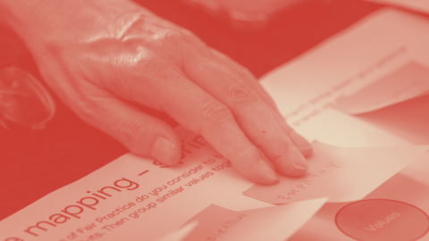 Close-up photo with a red monochrome duotone filter showing a hand placing a small paper note labeled "EMPATHY" onto a worksheet during a group exercise, likely a values-mapping or workshop activity. The visible worksheet reads "mapping" with instructions about grouping similar values together, and a circular sticker labeled "Values" is visible in the bottom right. A pair of glasses rests on the table in the blurred background.