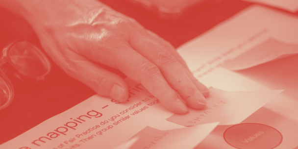 Close-up photo with a red monochrome duotone filter showing a hand placing a small paper note labeled "EMPATHY" onto a worksheet during a group exercise, likely a values-mapping or workshop activity. The visible worksheet reads "mapping" with instructions about grouping similar values together, and a circular sticker labeled "Values" is visible in the bottom right. A pair of glasses rests on the table in the blurred background.