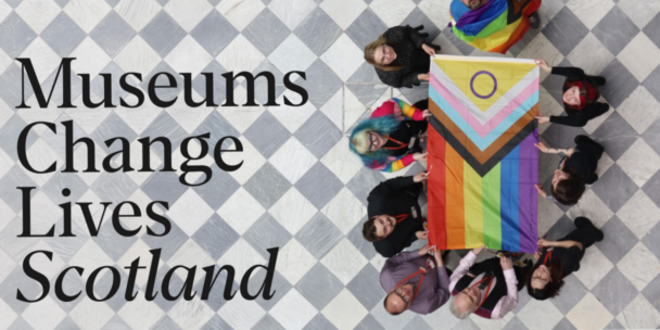 Overhead view of a group of people standing in a circle on a grey and white chequered marble floor, holding an intersex-inclusive Progress Pride flag between them. One person is wrapped in a second Pride flag. On the left, large black text reads "Museums Change Lives Scotland."