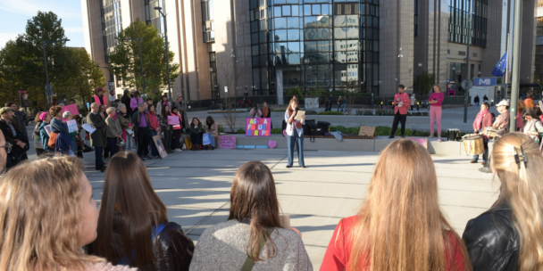 A picture of people attending the StandUpForAgoraEU protest in Brussels. An artist is delivering a performance in the centre, and people around are listening and carrying protest signs in front of the Council building.