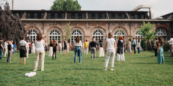 On a sunny day, a large group of people in light summer clothes stand spread out across a grassy courtyard, most with their backs to the camera. They face a long red-brick building with a row of tall arched windows. Young trees and a large dark-leaved tree frame the scene.